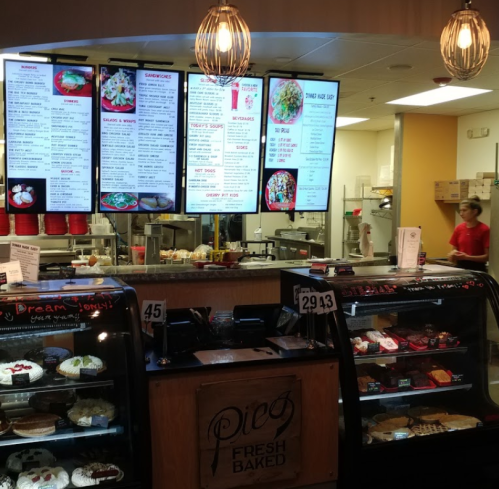 A bakery display with desserts and a menu board, featuring sandwiches and salads, in a cozy cafe setting.