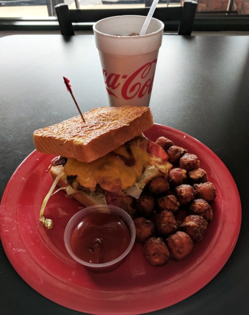 A sandwich with cheese, lettuce, and bacon on toasted bread, served with tater tots and a cup of soda.