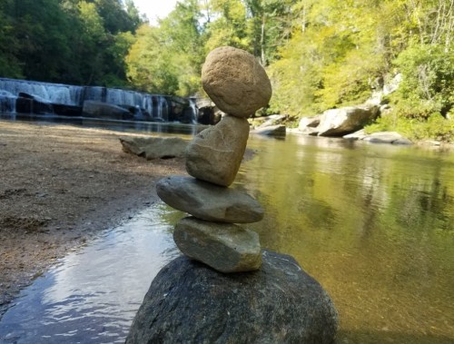 A stack of smooth stones balanced on a larger rock by a calm river, with a waterfall and trees in the background.