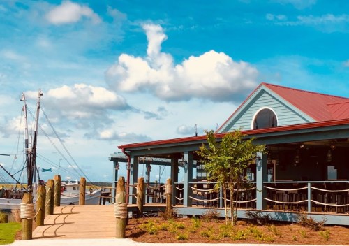 A waterfront restaurant with a red roof, surrounded by greenery and boats in the background under a blue sky.