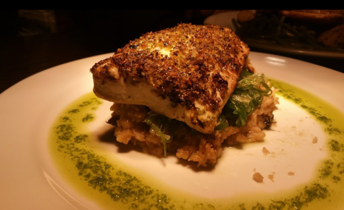 A beautifully plated dish featuring a crusted fish fillet atop a bed of grains and greens, drizzled with green sauce.