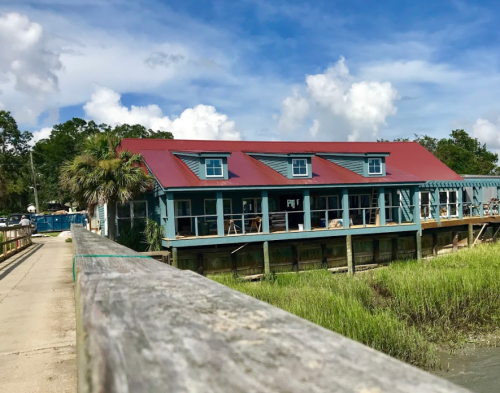 A waterfront building with a red roof and large windows, surrounded by greenery and a blue sky with clouds.