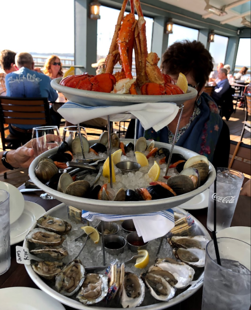 A seafood tower featuring crab, shrimp, clams, oysters, and lemon slices, set on a dining table with patrons in the background.