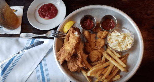 A metal tray with fried fish, shrimp, fries, coleslaw, and dipping sauces, alongside a plate of ketchup.