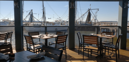 View of a waterfront restaurant with empty tables, boats docked in the background, and clear blue skies.