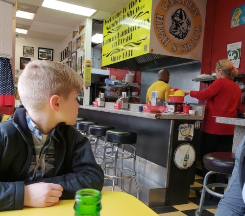 A young boy sits at a diner table, looking towards the counter where staff are preparing food. Retro decor surrounds them.