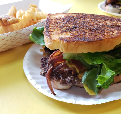 A close-up of a grilled sandwich with lettuce, bacon, and a beef patty, served with a side of fries in a basket.