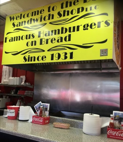 Bright yellow sign reading "Welcome to the D's Sandwich Shoppe" with a counter and condiment containers in a diner setting.