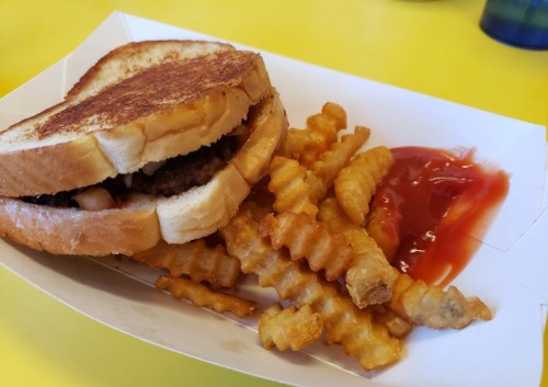 A grilled sandwich with a beef patty and crinkle-cut fries served with ketchup on a yellow table.