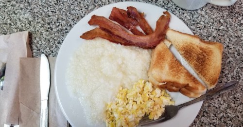 A plate of breakfast featuring scrambled eggs, grits, crispy bacon, and toasted bread on a speckled countertop.