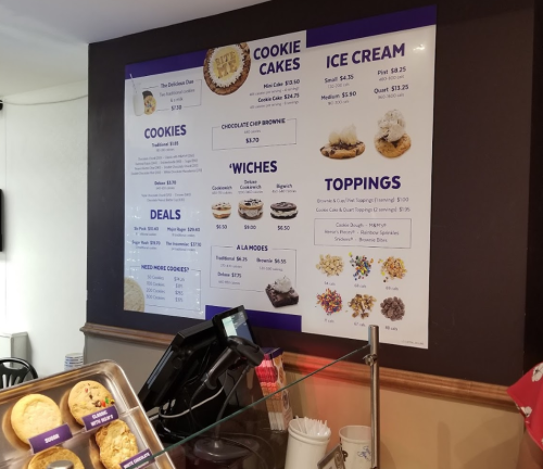 Menu board displaying cookie cakes, cookies, ice cream, deals, and toppings at a dessert shop.