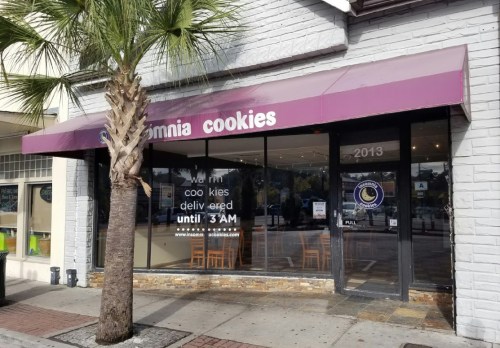 Exterior of a cookie shop with a purple awning, featuring a sign that says "Cookies delivered until 3 AM."