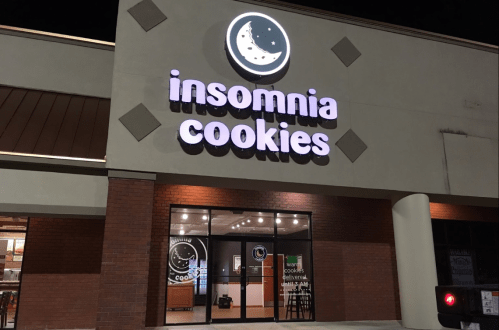 Exterior of Insomnia Cookies store at night, featuring a lit sign and large glass entrance.