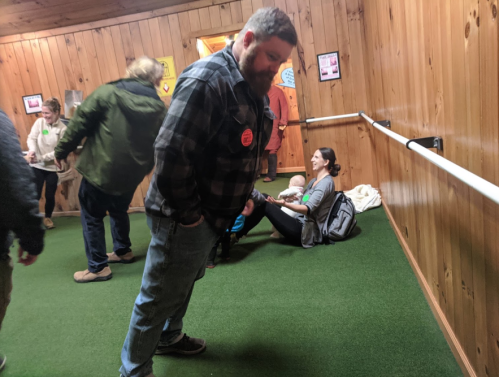 A man in a plaid shirt leans forward in a wooden hallway, while a woman sits on the floor with a baby nearby.