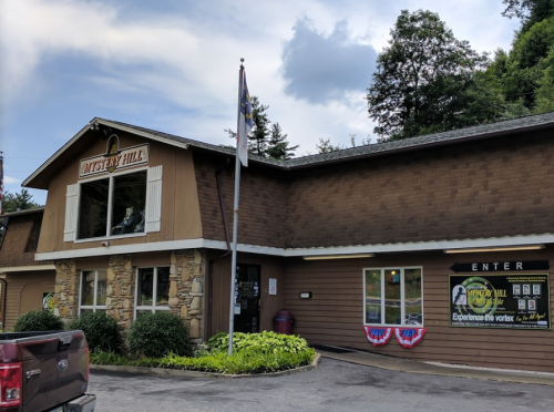 Exterior of Mystery Hill building with a flag, sign, and decorative plants under a cloudy sky.