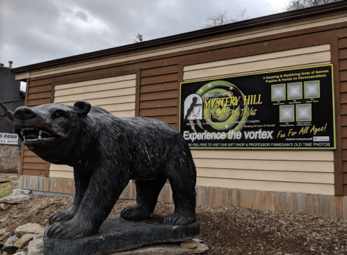 A bear statue in front of a sign for Mystery Hill, promoting games, puzzles, and vortex experiences.