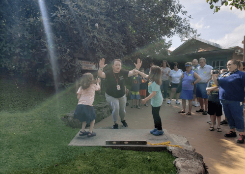 A group of people watches as a woman demonstrates an activity with two children outdoors. Sunlight filters through trees.