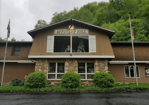 A brown building with a stone facade, featuring a sign that reads "Mystery Hill" and an open flag, surrounded by greenery.