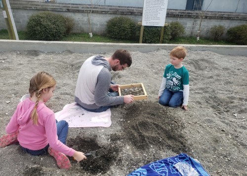 A man and two children sift through gravel in a sandy area, engaged in a digging activity.