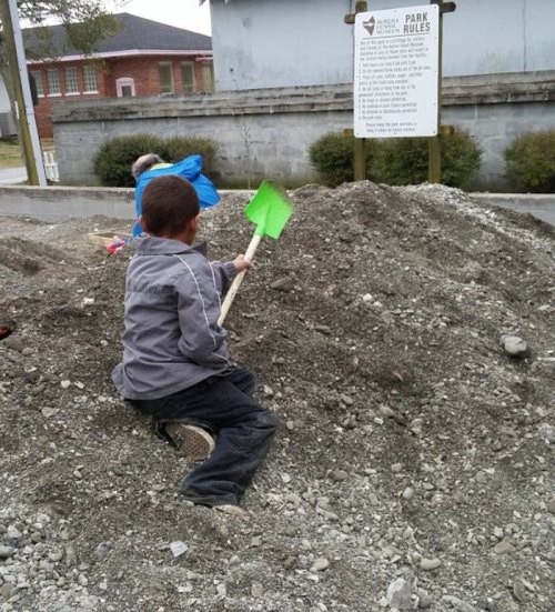 A child sits on a dirt pile, holding a green shovel, while another child plays nearby. A park rules sign is in the background.