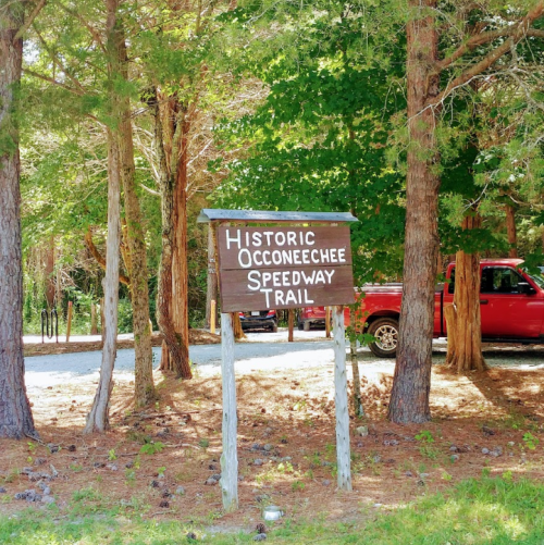 Sign for the Historic Occoneechee Speedway Trail surrounded by trees, with a red truck parked nearby.