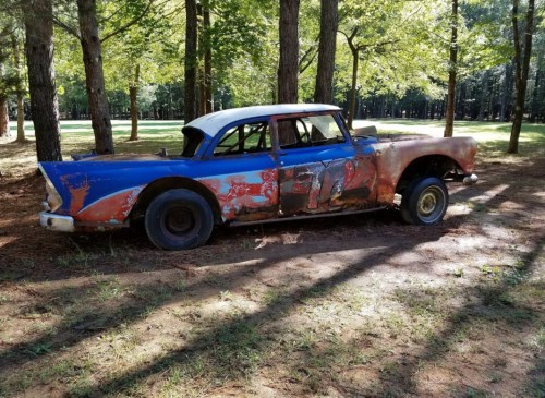 An old, rusty car with a blue and orange paint job, parked among trees in a grassy area.
