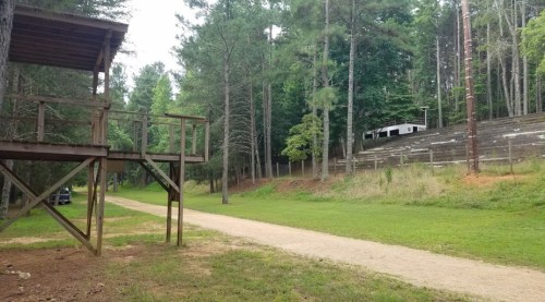 A dirt path winds through a forested area with a wooden structure on the left and a building on a hill in the background.