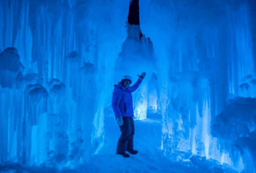 A person in a purple jacket waves inside a glowing blue ice cave with icicles hanging from the walls.