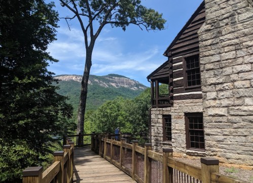 A wooden walkway leads to a stone building, with a mountain and blue sky in the background. Lush greenery surrounds the scene.
