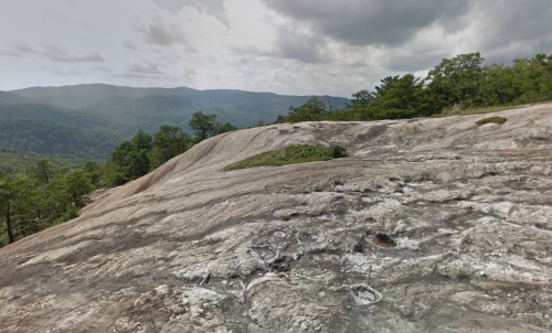 A rocky landscape with a smooth surface, surrounded by green trees and distant mountains under a cloudy sky.