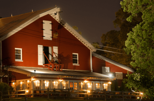 A red barn at night, illuminated by warm lights, surrounded by trees and power lines.