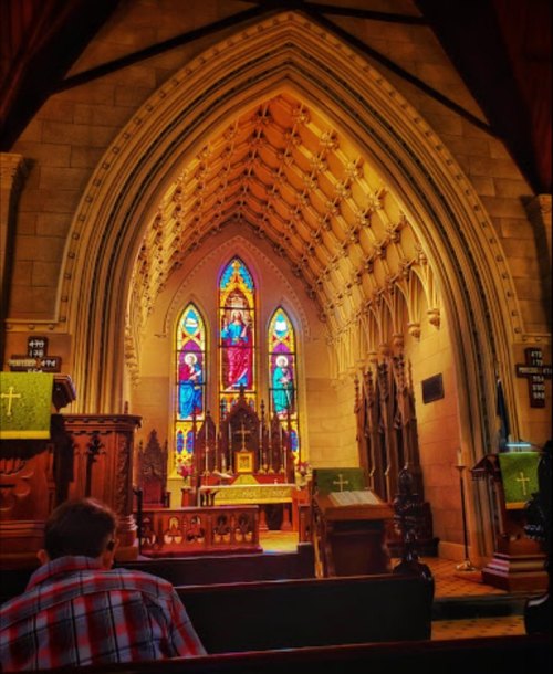 Interior of a church featuring a colorful stained glass window and ornate architectural details, with a person seated in prayer.