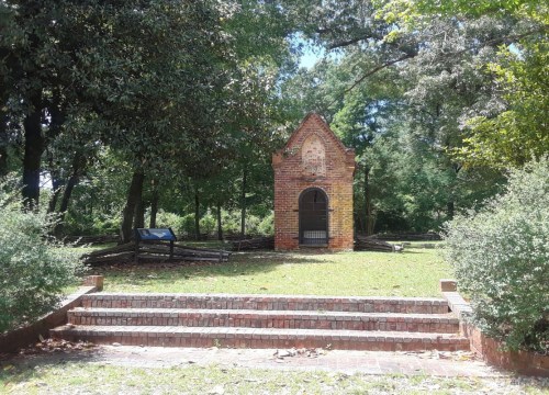 A small brick structure surrounded by trees, with steps leading up to it and a sign nearby.