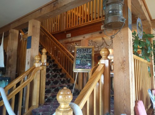 Interior of a rustic restaurant with wooden stairs, a chalkboard sign, and decorative lights.