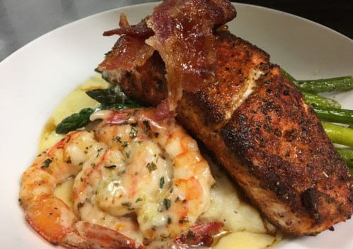 A plate featuring seasoned salmon, shrimp, asparagus, and creamy mashed potatoes, garnished with crispy bacon.