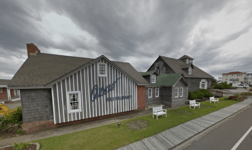 A rustic restaurant with a striped exterior and a green roof, surrounded by benches and a cloudy sky.