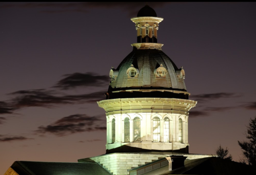 A historic building's dome illuminated at night, with a cloudy sky in the background.