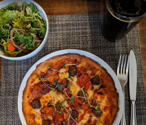 A plate of pizza topped with sausage and herbs, alongside a fresh salad and a glass of dark beverage on a textured placemat.
