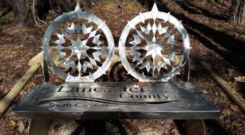 A metal bench featuring star designs and engraved text for Lancaster County, South Carolina, set in a wooded area.