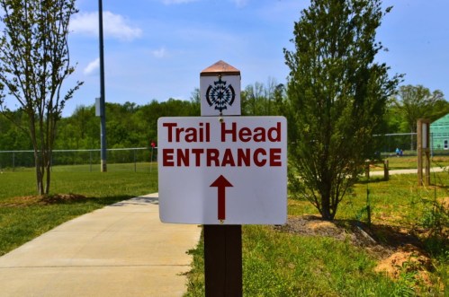 Sign indicating the entrance to a trail head, with an arrow pointing upward, surrounded by greenery.
