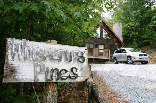 A wooden sign reading "Whispering Pines" near a cabin and a parked car on a gravel road surrounded by trees.