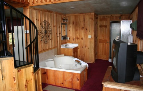 Cozy wooden interior featuring a spiral staircase, a jacuzzi tub, and a TV in a rustic cabin setting.