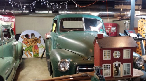 A vintage green truck in a cluttered garage with a small dollhouse and a colorful mural of dancers in the background.