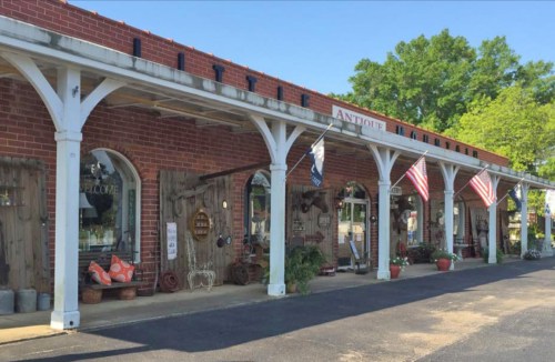 A charming storefront with wooden accents, American flags, and potted plants, set against a clear blue sky.