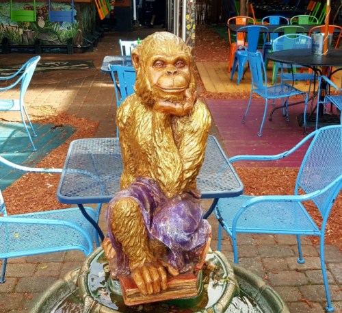 A golden monkey statue sitting on books, resting its chin on one hand, surrounded by colorful outdoor seating.