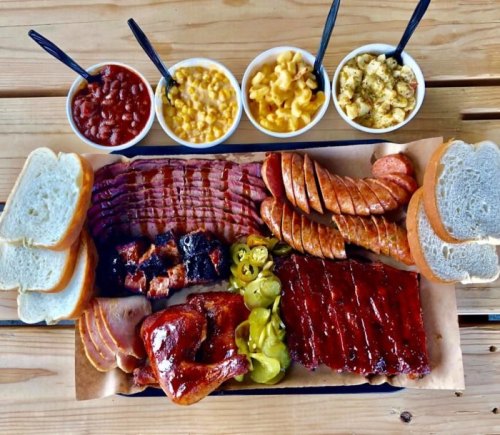 A platter of assorted barbecue meats, including ribs, sausage, and chicken, served with sides of beans, mac and cheese, and bread.