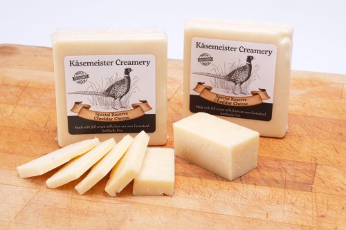 Two blocks of Käsemeister Creamery cheddar cheese on a wooden cutting board, with slices arranged beside them.