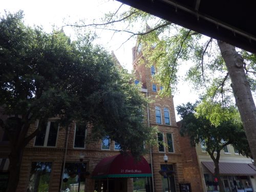 Historic brick building with a clock tower, surrounded by trees and shops, under a clear blue sky.