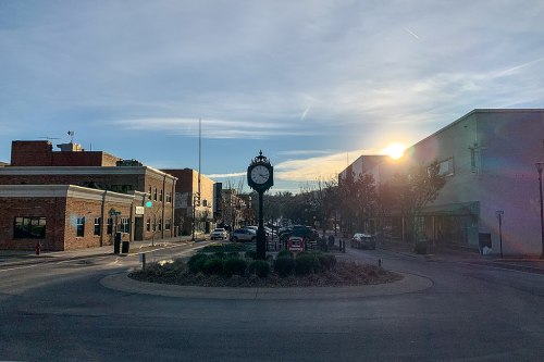 A clock tower in a town square at sunset, surrounded by buildings and trees, with cars parked along the street.