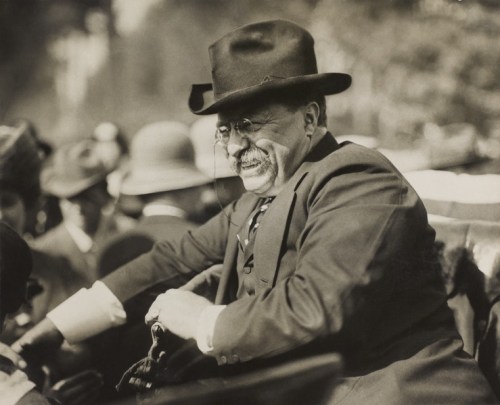 A historical black-and-white photo of a smiling man in a hat, seated and engaging with a crowd.